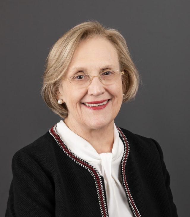 A headshot of an older white woman with a blonde bob haircut. She's wearing glasses and a soft white blouse covered by a black blazer with a threaded white, red, and black trim. 