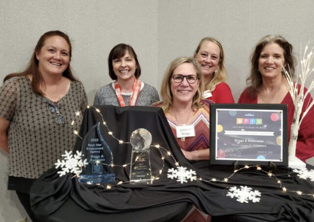 Group picture of 5 women from Briggs and Associates standing behind a table displaying award plaques and trophies that they have received.