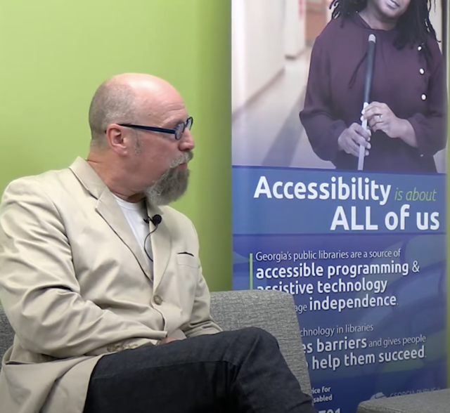 Photo of Doug Crandell’s profile while he sits in a chair. A poster is behind him with the words Accessibility for All.