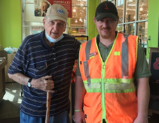 Photo taken in a restaurant of Thomas Fewell, a white male dressed in a bright orange safety shirt with yellow reflectors and has a ball cap on, standing to the right of his friend Buddy, who is an elderly male with a ball cap and a blue and white striped shirt while holding his walking cane