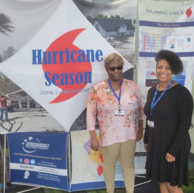Foto de Naomi y otra señora afroamericana paradas frente a una pancarta que dice Temporada de huracanes