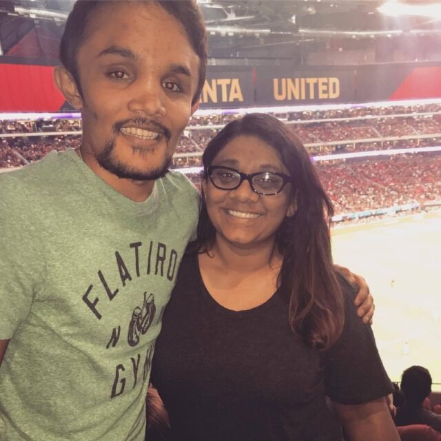 A tall Indian man wearing a grey t-shirt smiles with a short Indian woman wearing a black t-shirt and glasses. The background is a sports stadium. 