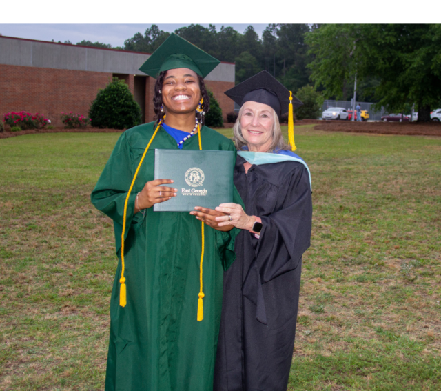 A photo of a young African American female graduate holding her diploma and and wearing a green graduation tam, gown, and hood, while standing to the left of an older white woman wearing a black graduation tam, black gown, and blue hood.