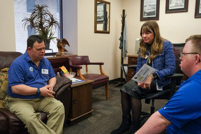 two advocates wearing blue t-shirts speak to a legislator in an office