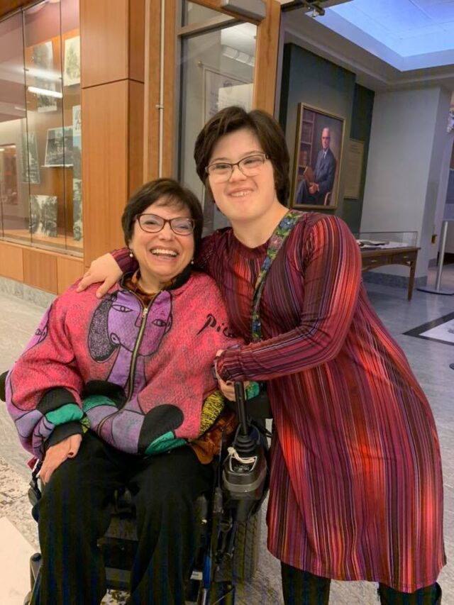 Two women pose for the camera smiling. The woman on the left is in a wheelchair and wearing a pink jacket and glasses. The woman on the right is standing and hugging the other women wearing a red dress and glasses. 