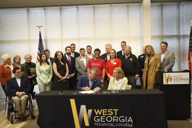 Governor Brian Kemp sitting at a large brown desk signing the bill HB185 with a large group of Destination Dawgs and GCDD representatives behind him.