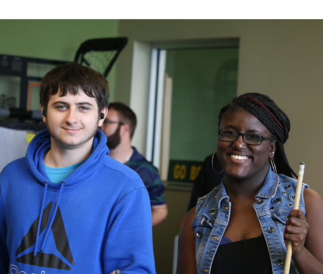 A photo of a young white man wearing a blue hoodie while smiling and a young smiling black woman wearing glasses and a black tank top under a blue jean vest while holding a pool table stick