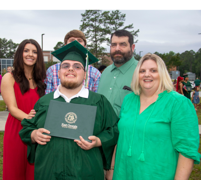 A photo of a caucasian family including a young white woman wearing a red dress, a young brunette white man wearing a colorful plaid shirt, an older white brunette man wearing a green dress shirt, an older white, blonde woman wearing a green blouse with tassels, and a young white brunette man in the center wearing a green graduation cap and gown