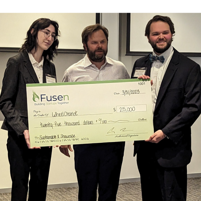 Stoppard and Jones, both wearing black business suits, holding a large check for $25,000 from the company FUSEN with a business man standing between them