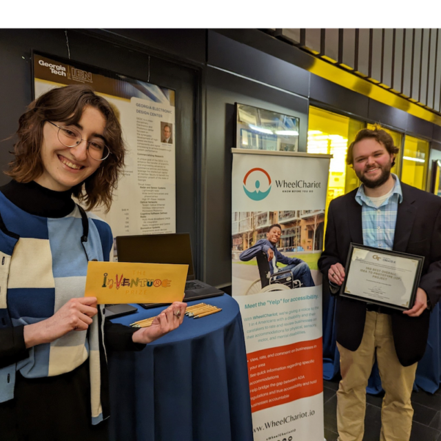 This is a picture with a young white, brunette woman on the left side holding a piece of paper that says "inventure" and a young white, brunette man on the right side of the picture holding an award certificate. In the middle of the two people is a retractable banner with Wheelchariot's logo on the back.