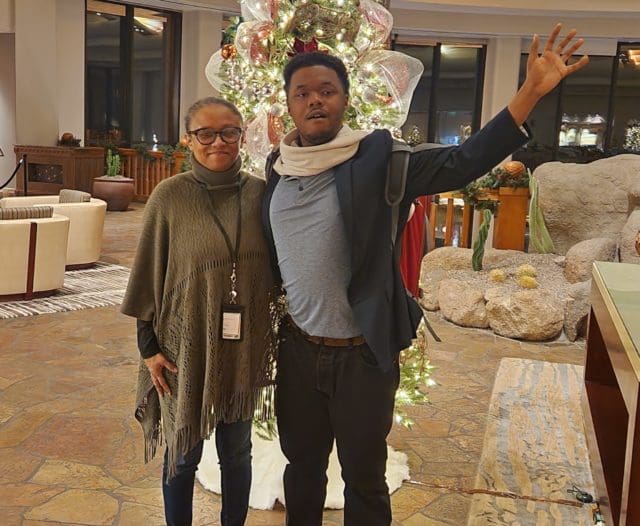A photo of an African American woman wearing glasses and a muted green shawl standing next to her African American young adult son, whose wearing a gray shirt, black blazer, and beige scarf